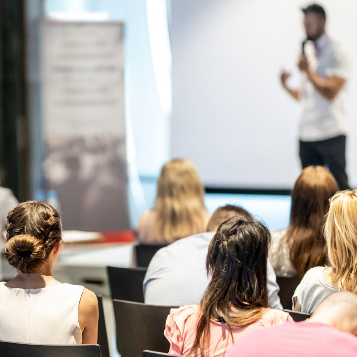 photo of people seated listening to a person speaking while holding a microphone